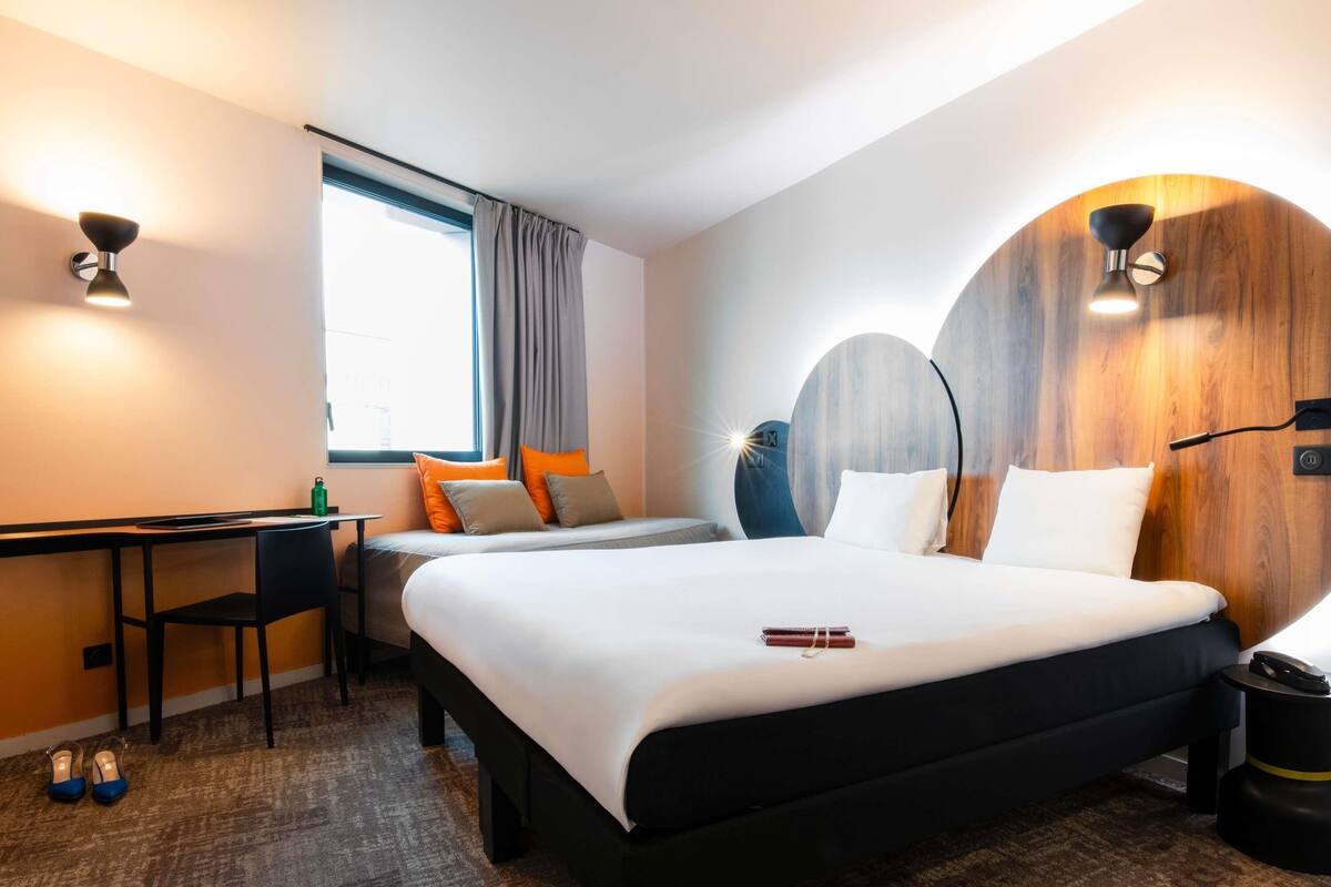Une chambre d’hôtel comprenant un lit, un bureau, une chaise, une fenêtre avec des rideaux et un luminaire fixé au mur.