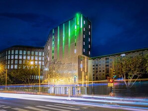 A modern building with a distinctive green-lit facade, a visible entrance, and a clear view of the surrounding urban environment.