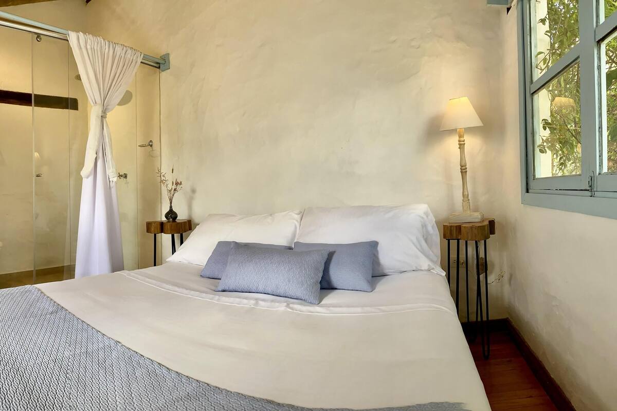 A bedroom with a bed, bedside tables, a window, and a bathroom visible through an open door.