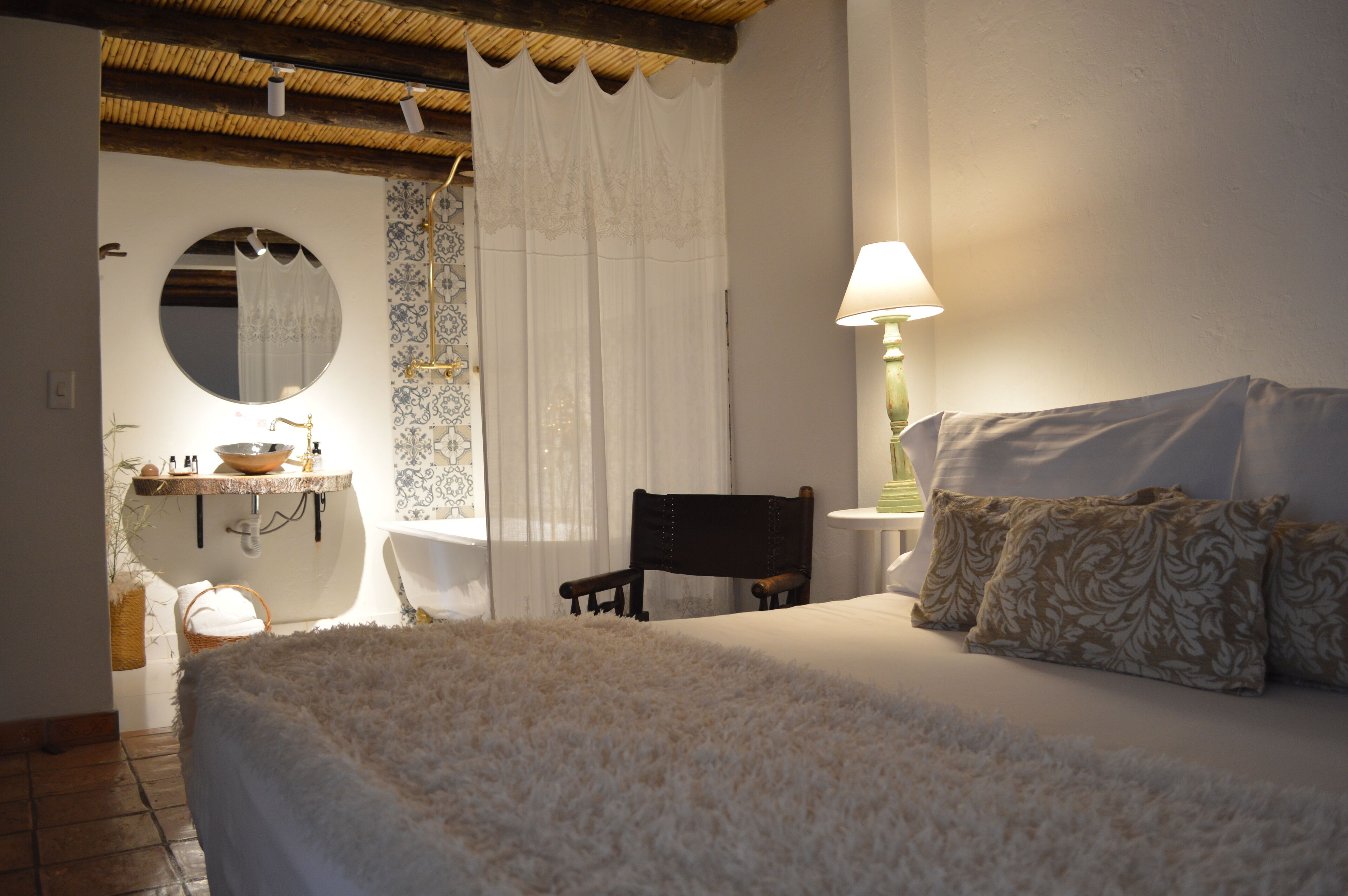 Un dormitorio con cama, una silla, una mesita de noche con lámpara, un espejo y un baño con lavabo y toallero.