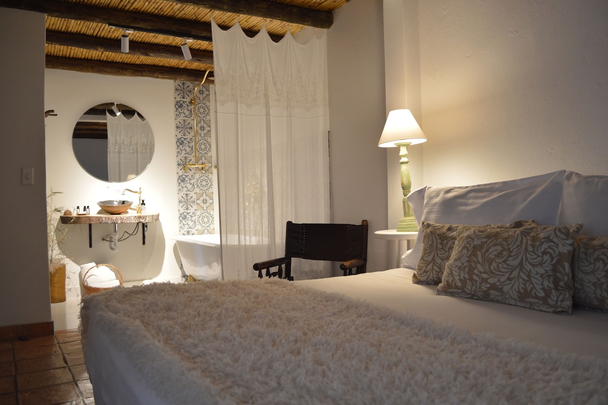 A bedroom with a bed, a chair, a bedside table with a lamp, a mirror, and a bathroom with a sink and a towel rack.