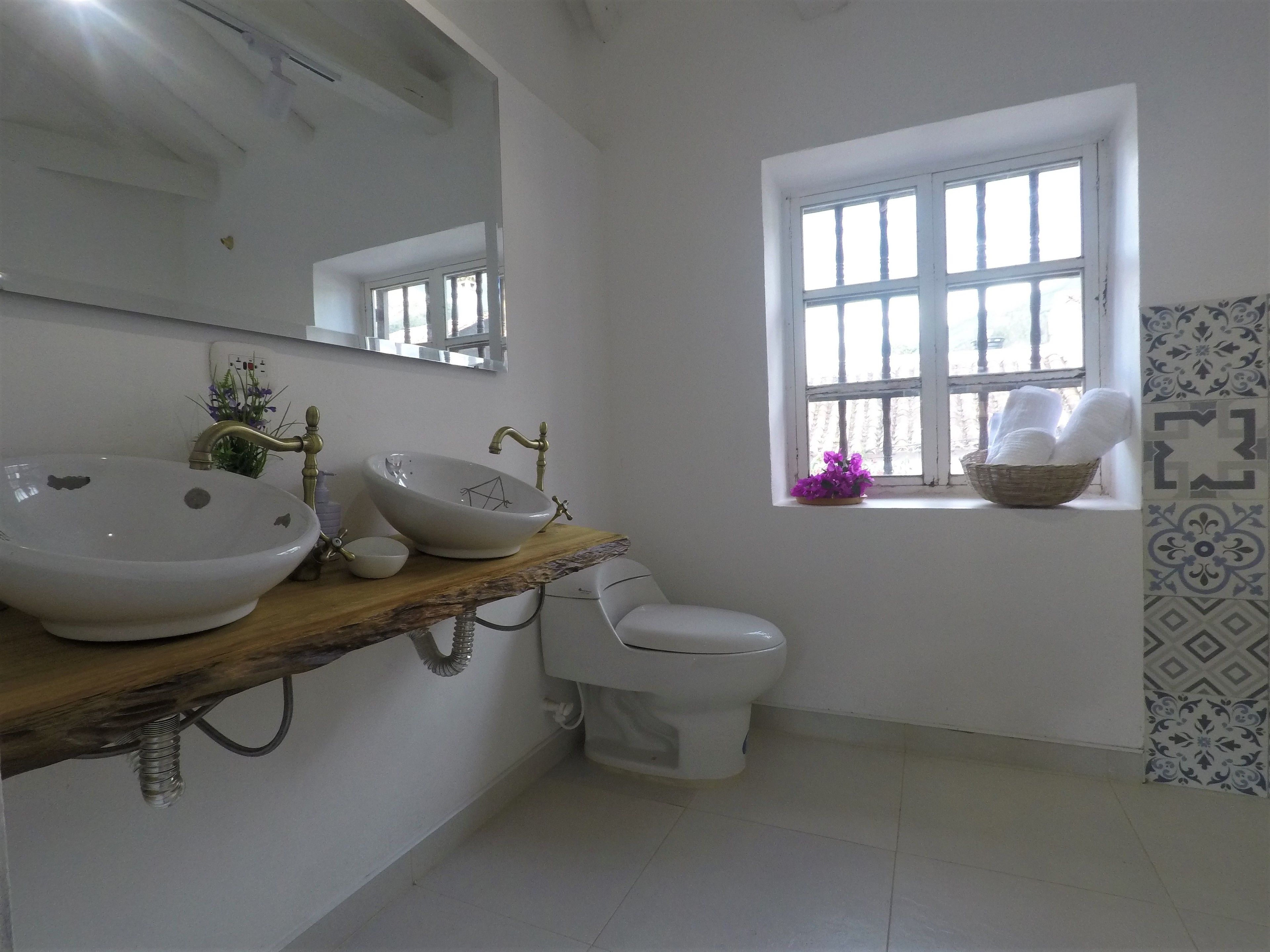 Un baño con dos lavamanos, un inodoro, una ventana con una cesta de toallas y un espejo.