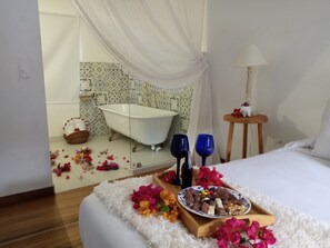 Un dormitorio con cama, una mesita de noche con lámpara, una bandeja con aperitivos y flores, y un baño con bañera y ducha.