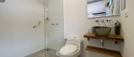 Un baño moderno con ducha envuelta en vidrio, inodoro blanco y tocador de madera con un lavabo grande.