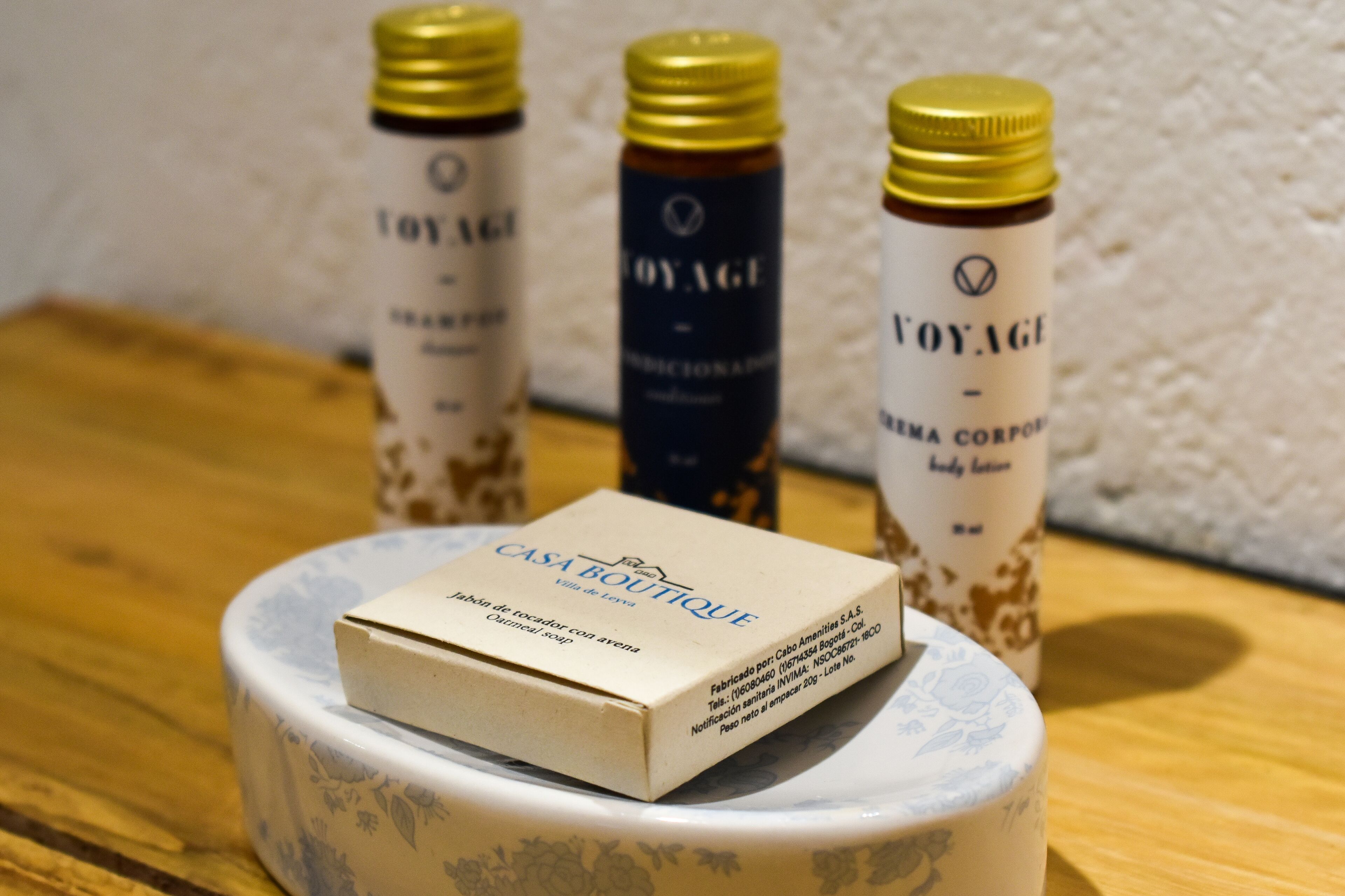 Una scatola con l'etichetta "CASA BOUTIQUE" su un elegante supporto in ceramica, con tre flaconi di lozione corpo "VOYAGE" sullo sfondo.