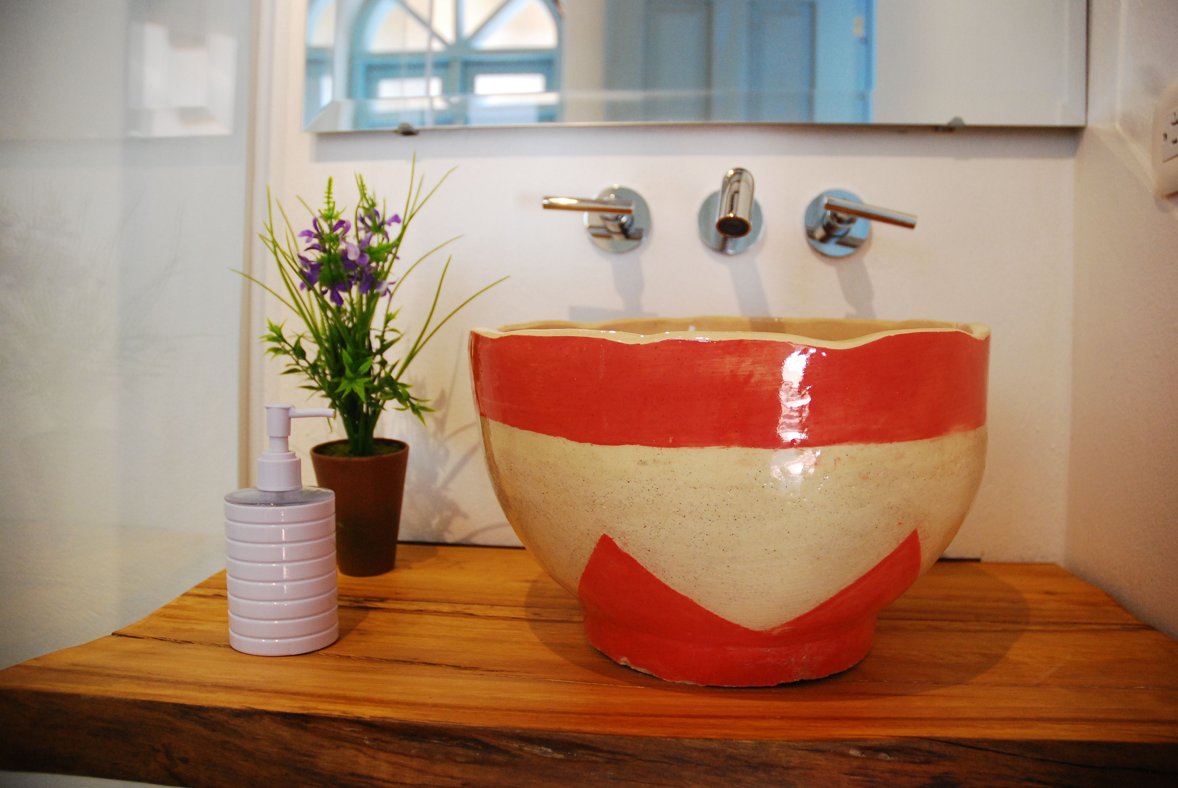 Une salle de bain avec un grand lavabo en céramique rouge et blanc, une plante en pot et un distributeur de savon posés sur un comptoir en bois.