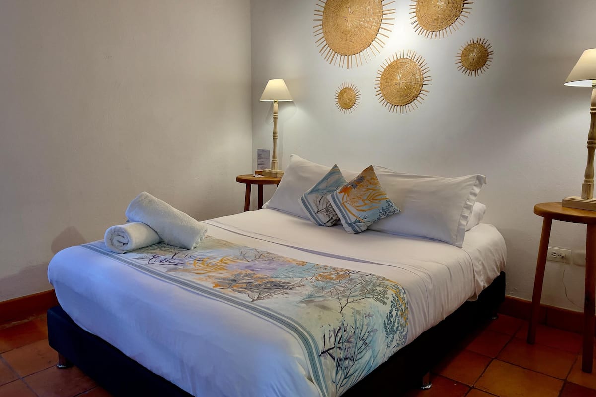 A neatly made bed with a floral bedspread, two pillows, and a rolled towel. There are two bedside tables with lamps, and wall art above the bed.