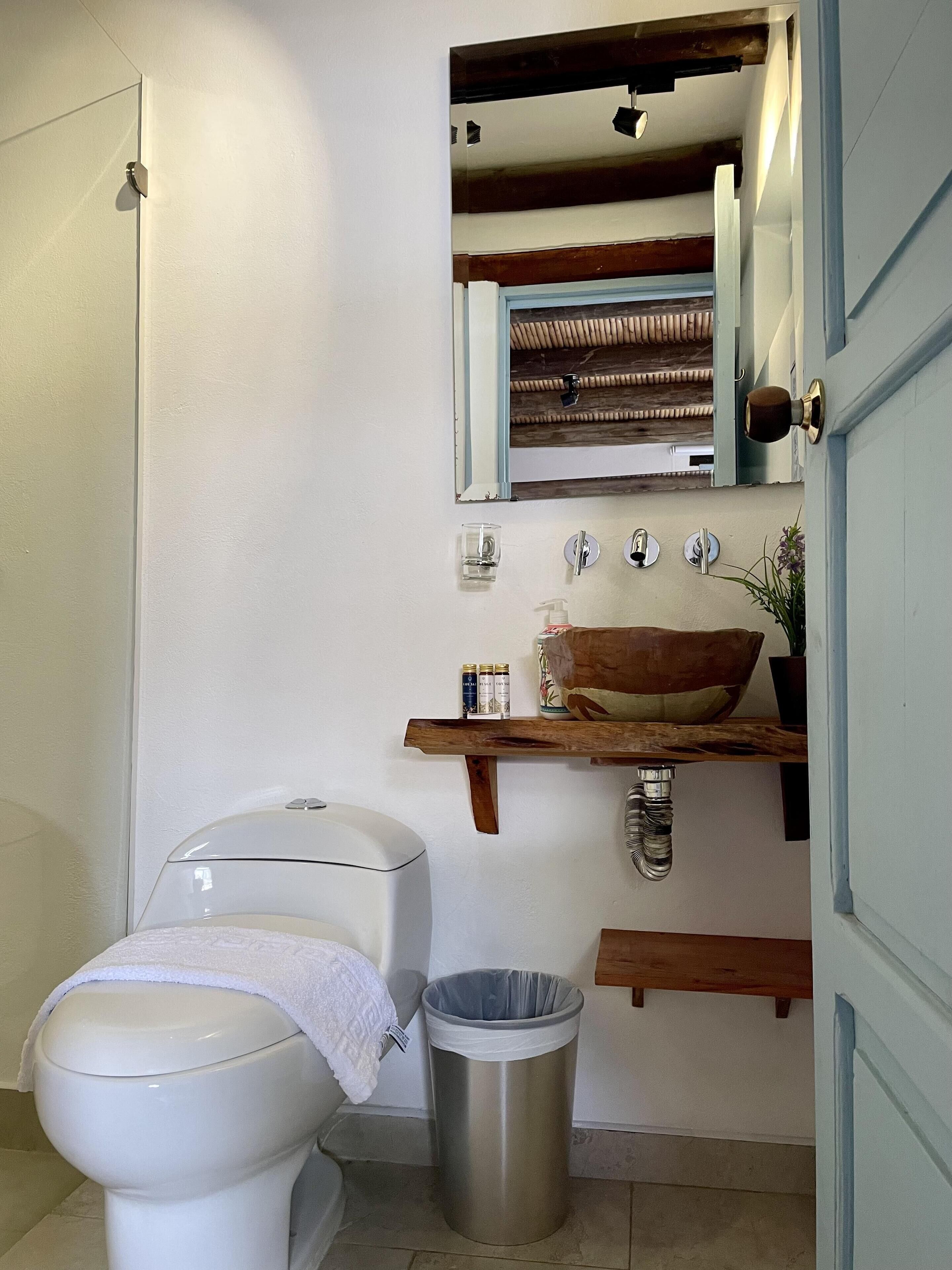 Un baño con inodoro, lavamanos con estante de madera, cesto de basura y espejo sobre el lavamanos.