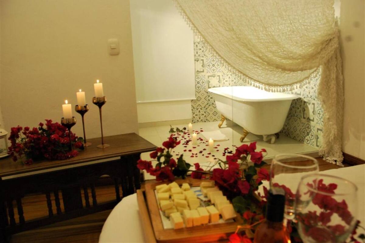 A room with a bathtub, candles, and a table with flowers and cheese.