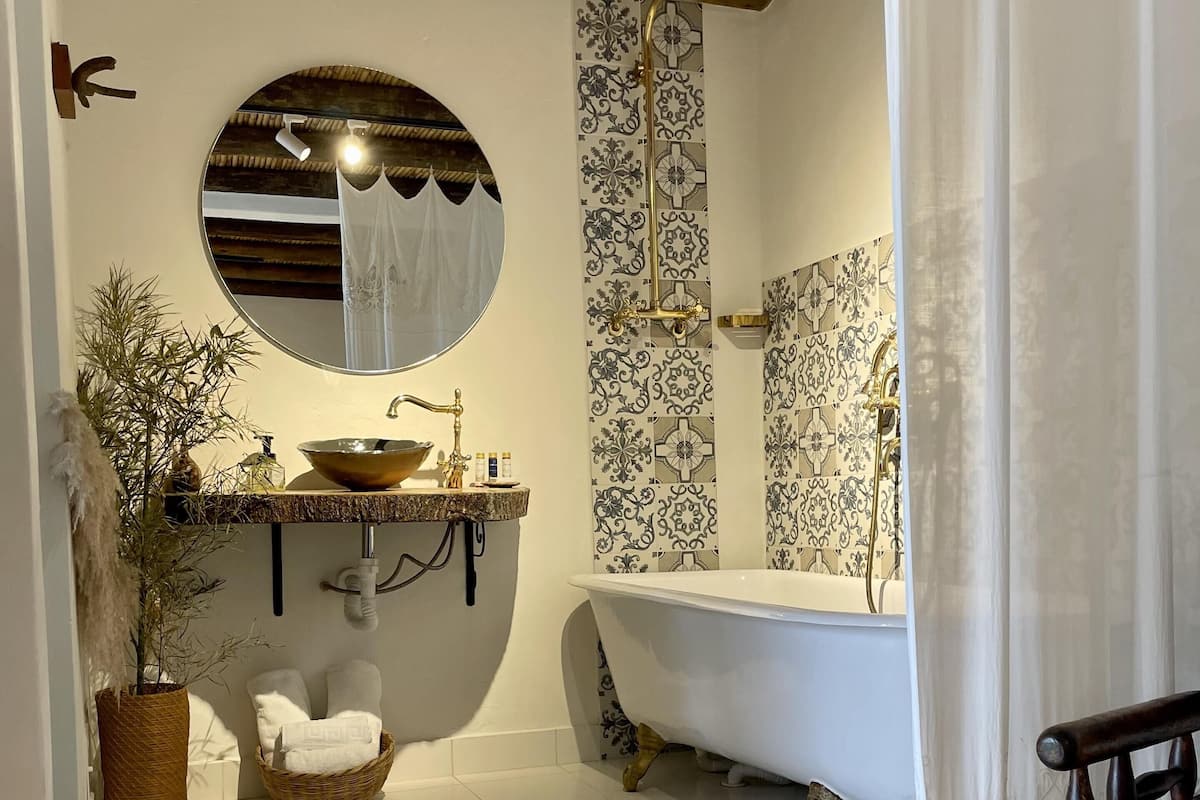 A bathroom with a freestanding bathtub, a round mirror, a sink with a decorative bowl, patterned tiles, and a bamboo ceiling.