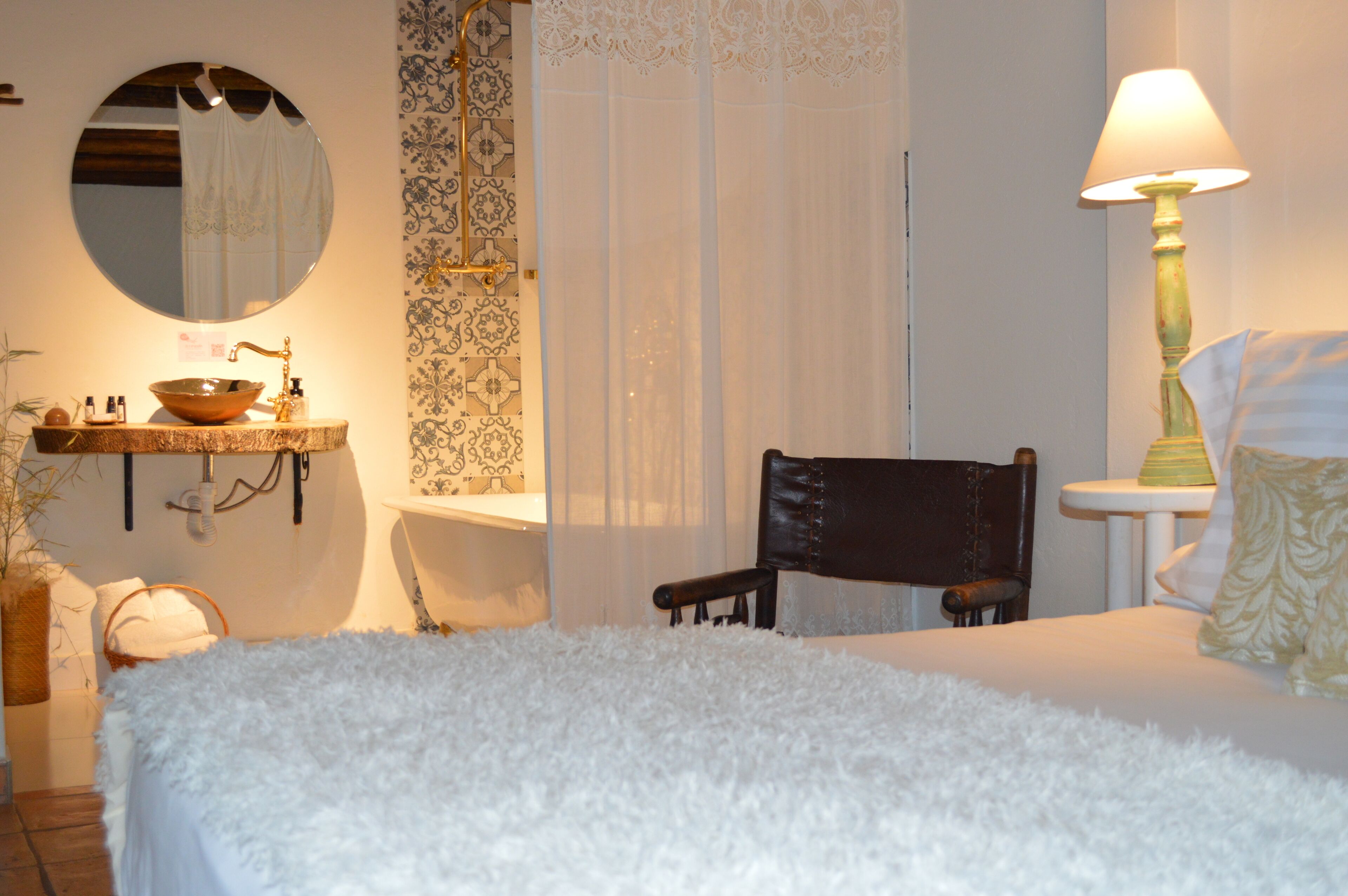 Quarto com espelho redondo, cadeira de madeira, cama com cobertura branca e fofa, mesa de cabeceira com abajur e banheiro com pia e espelho.