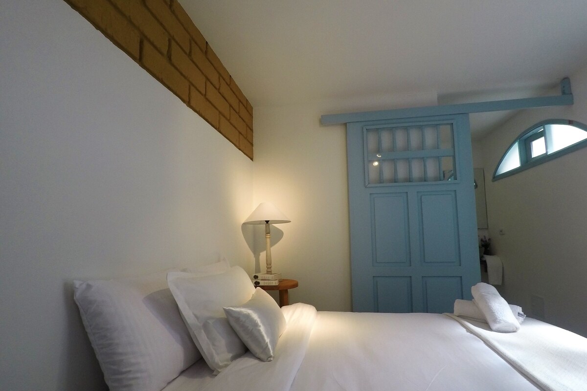 Una cama bien hecha con sábanas y almohadas blancas, una lámpara de noche y una puerta azul con un ventanal.