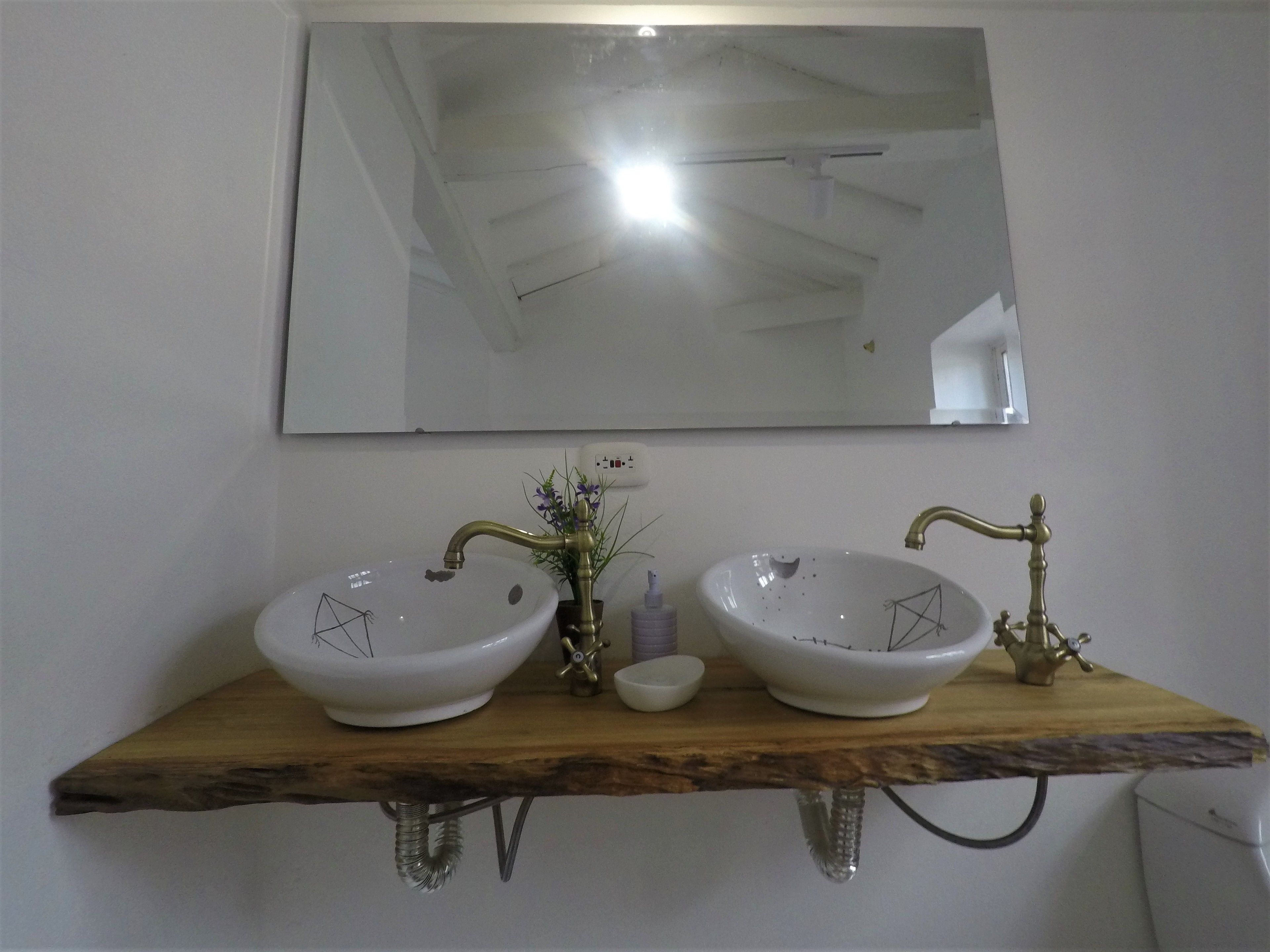 Un baño con dos lavamanos blancos, un estante de madera, un espejo y un inodoro.