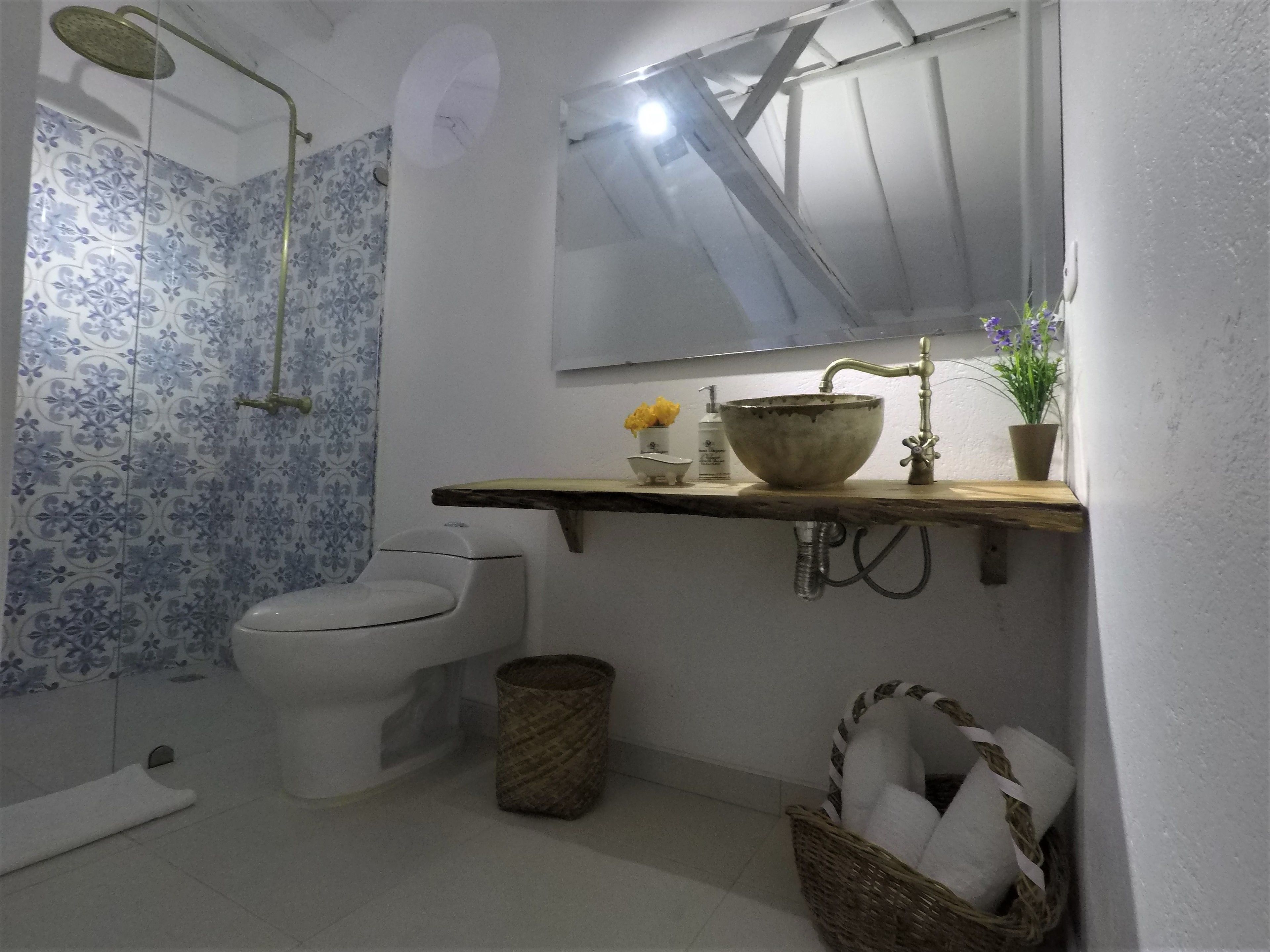 Un baño con inodoro, ducha, lavabo y espejo encima, y una pared con azulejos decorados.