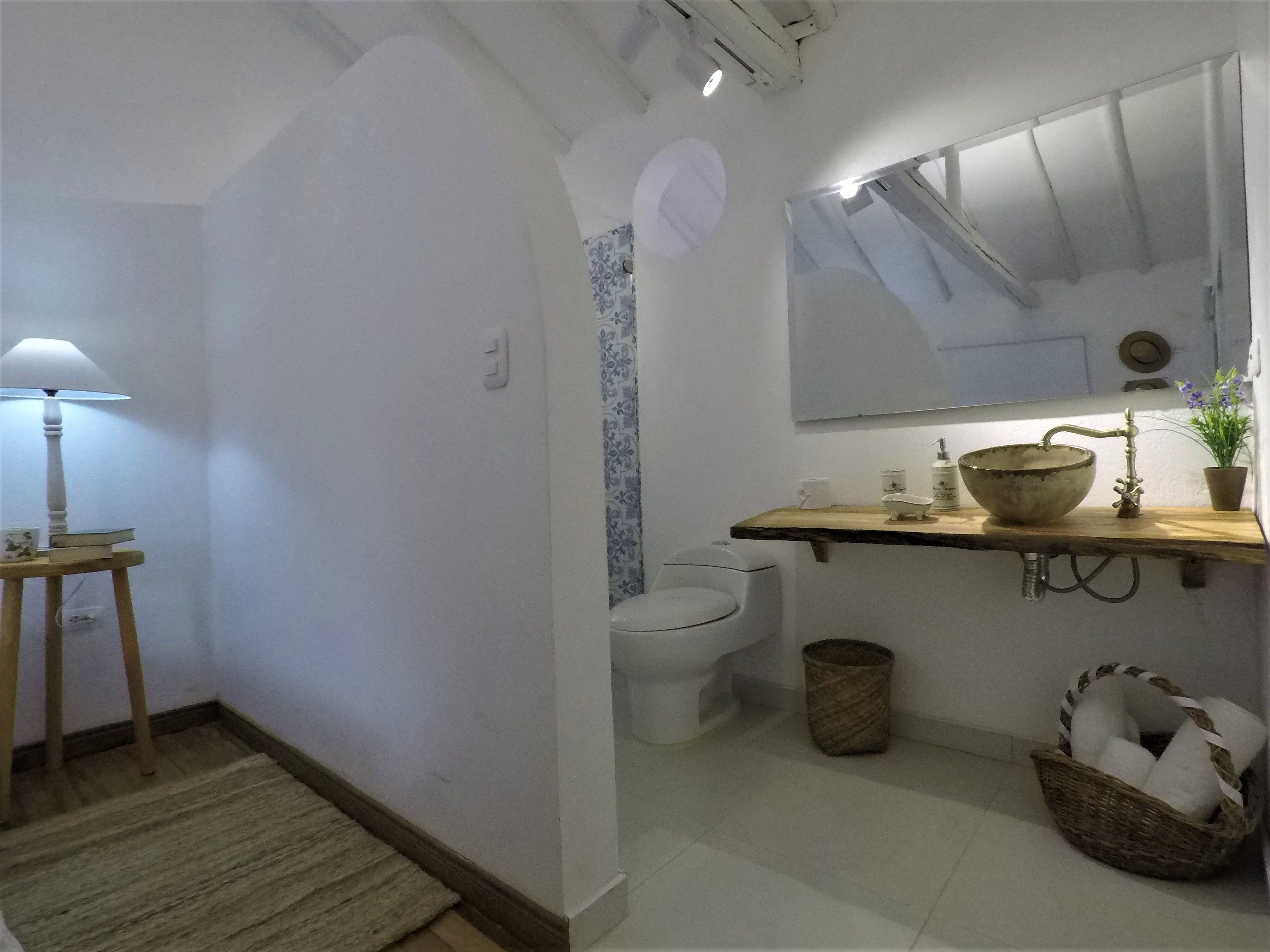 A bathroom with a white toilet, a wooden shelf with a basin and toiletries, a mirror, and a lamp on a side table.