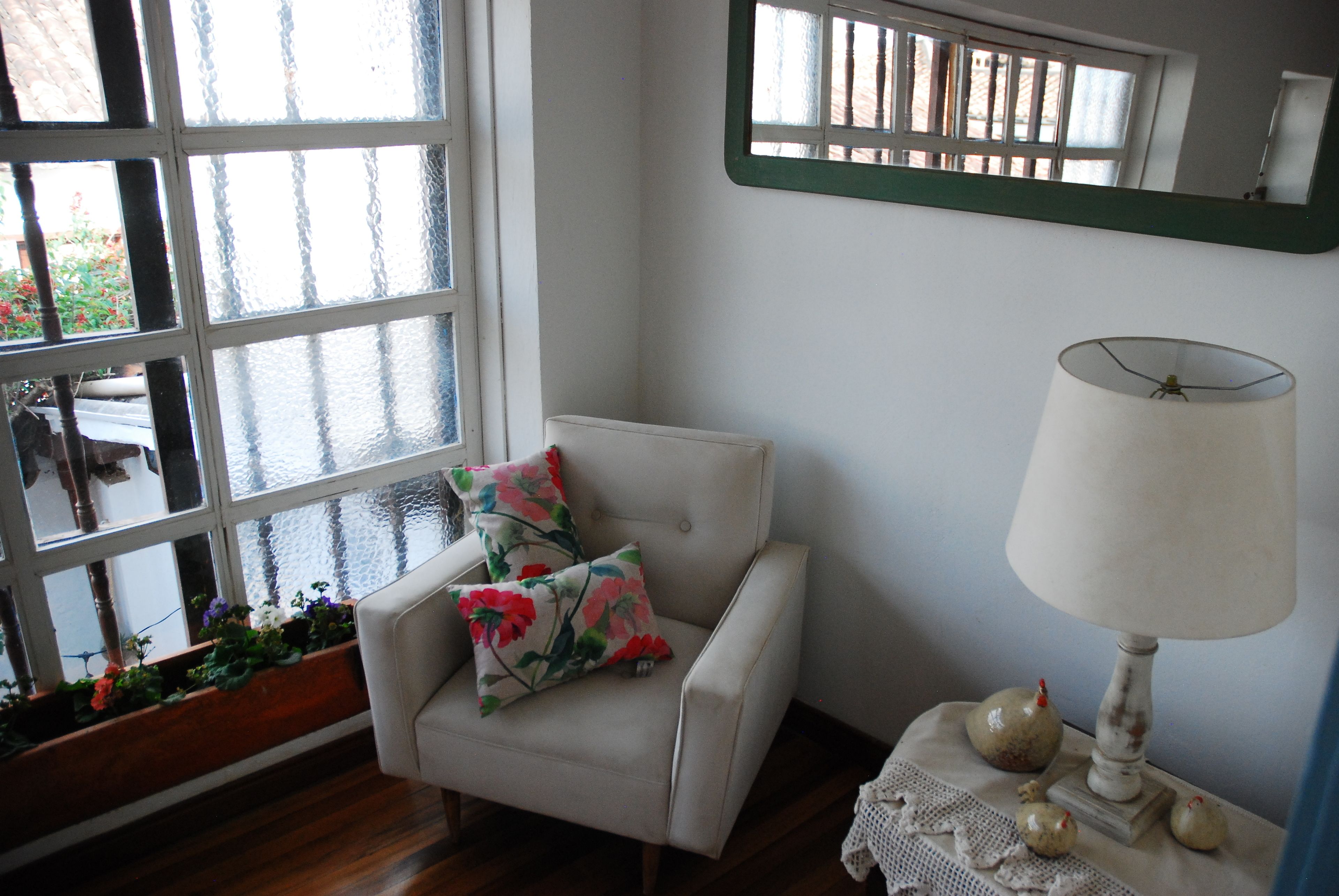 A room with a white armchair, a table with a lamp, and a window with a view of greenery outside.