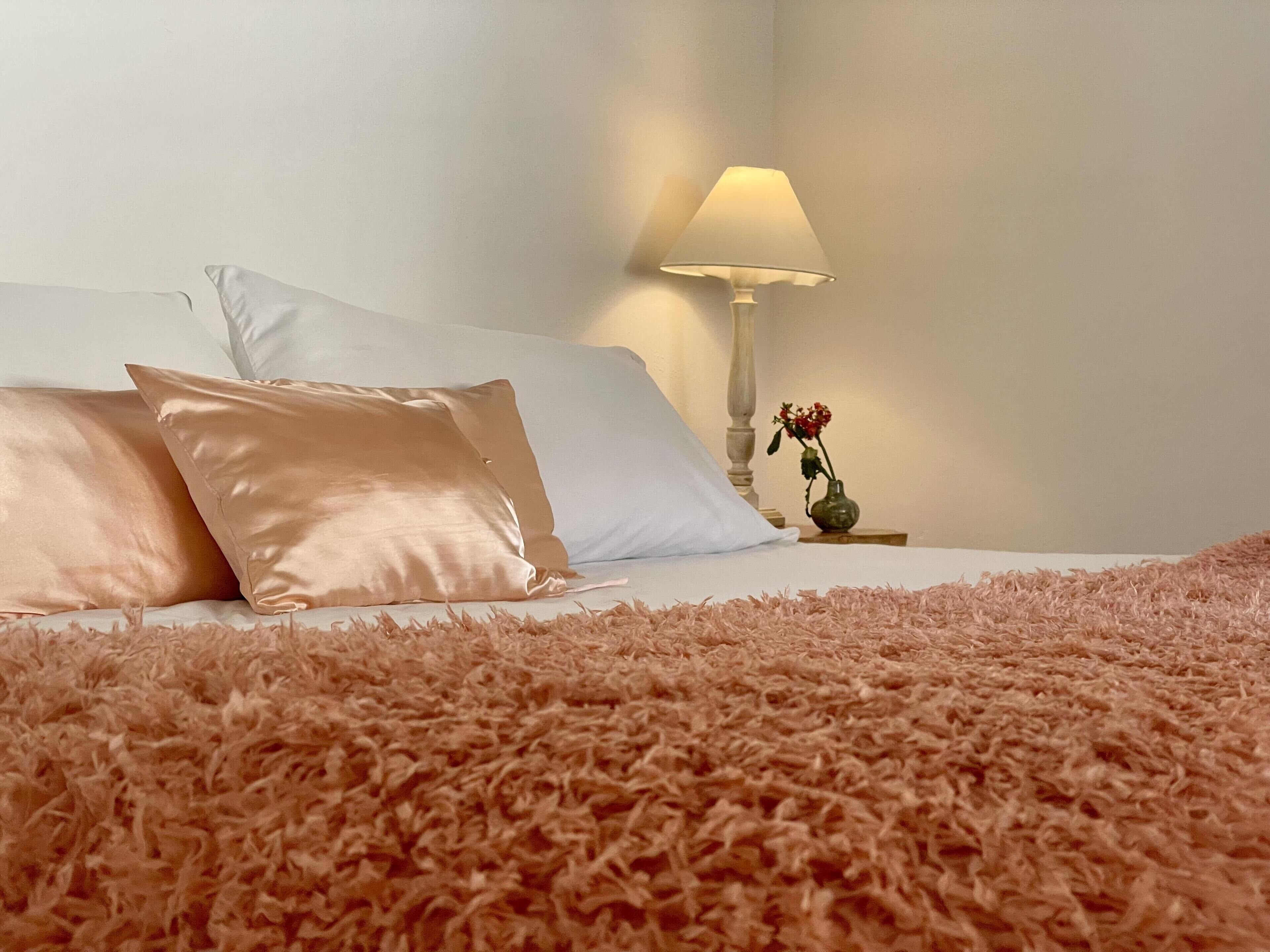A bed with a fluffy pink blanket, white and pink pillows, and a bedside table with a lamp and a vase of flowers.