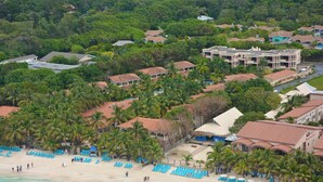 An aerial view of a coastal resort with multiple buildings, a beach with blue lounge chairs, and palm trees. - MAYAN PRINCESS BEACH AND DIVE RESORT (ROATAN)