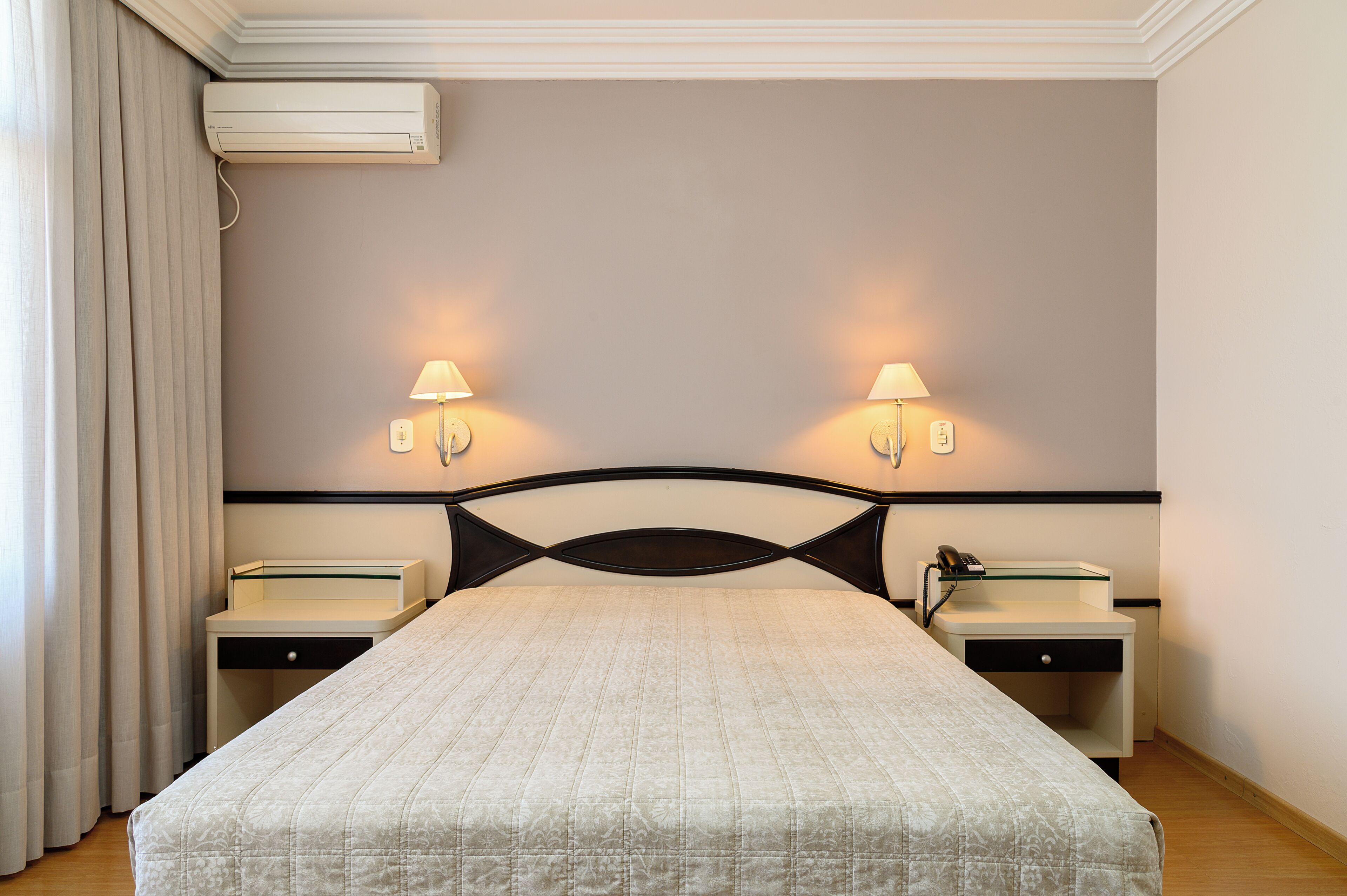 A hotel room with a bed, two bedside tables, a lamp, an air conditioner, and a telephone.