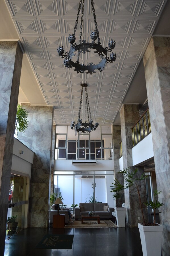 A spacious lobby with a high ceiling, marble walls, and a chandelier. There is a seating area with a sofa and armchairs, and potted plants are placed around the room. - Hotel Maerkli (Santo Angelo)