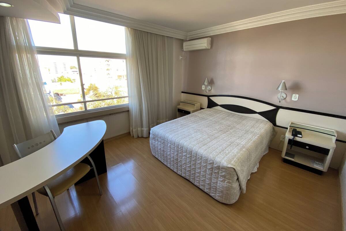 Ein Hotelzimmer mit Bett, Schreibtisch, Stuhl, Klimaanlage und einem fensterbepflanzten Fenster.