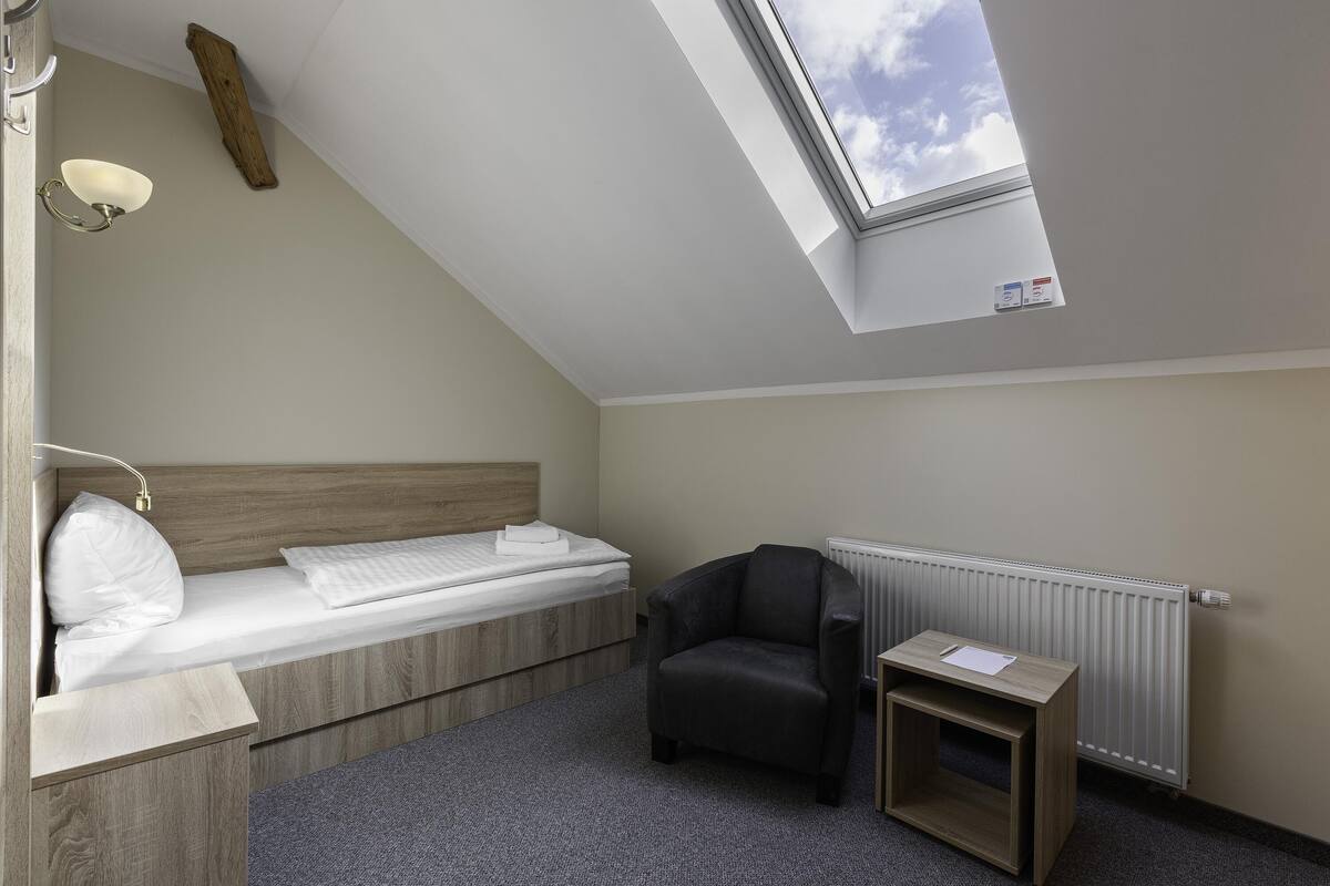 A small, modern room with a sloped ceiling, a bed, a chair, a small table, and a skylight.