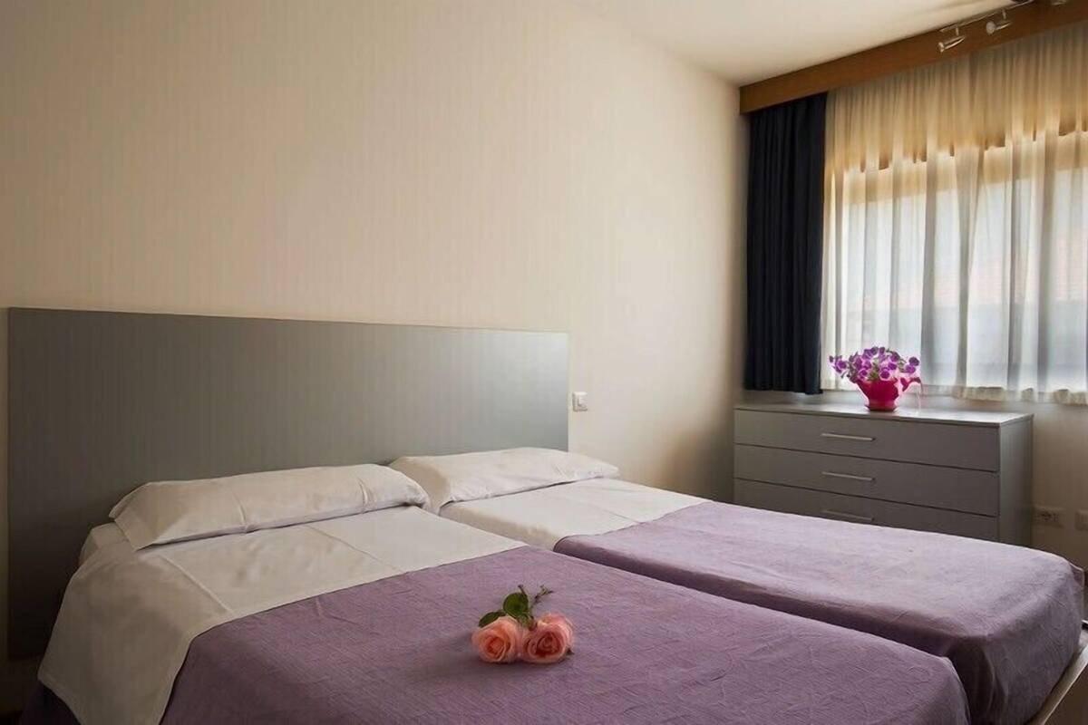A hotel room with two beds, a bedside table with a vase of flowers, and a window with sheer curtains.