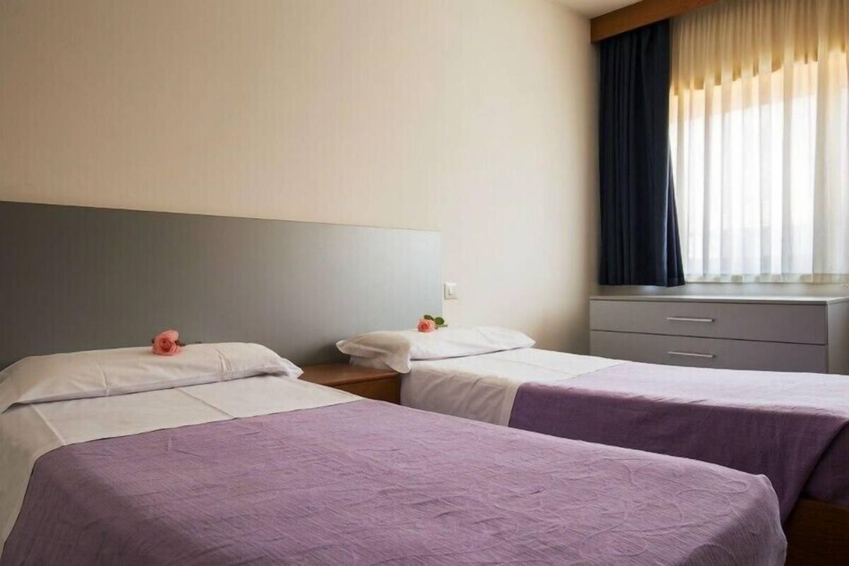 A hotel room with two beds, a nightstand, a dresser, and a window with curtains.