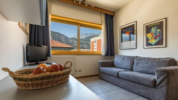 A modern living room with a grey sofa, a television, a wicker basket with fruits, and a window with a mountain view. - Residence Capitol (Trento)