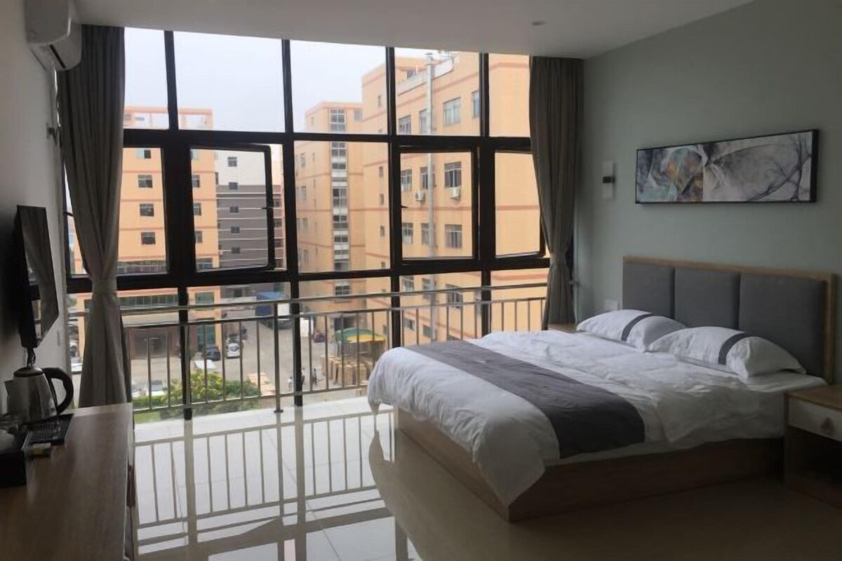 A bedroom with a large window, a bed, a nightstand, a painting, and a view of buildings outside.