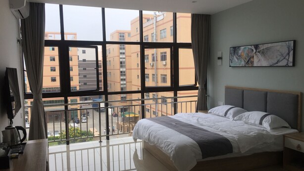 A hotel room with a large window, a bed, a bedside table, and a painting on the wall. - First Apartment (Jiangmen)