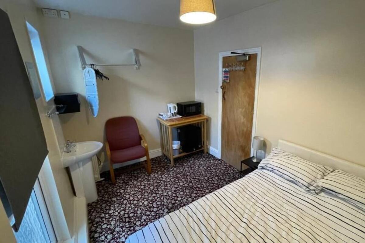 A small, compact room with a bed, a red chair, a small table with a microwave, a sink, and a door with a sign.