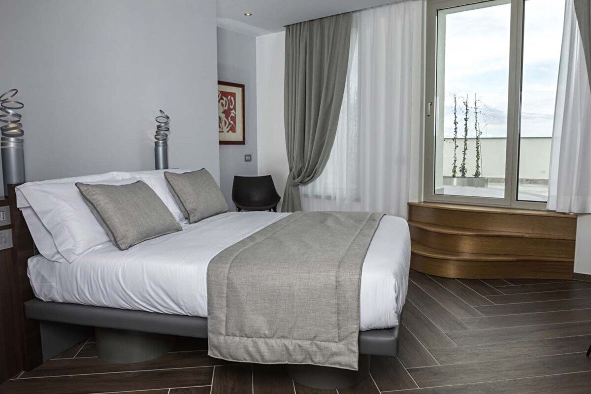 A modern bedroom with a bed, a chair, a window with curtains, and a view of the outdoors.