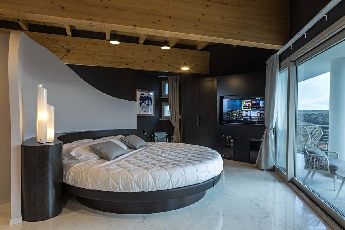 A modern bedroom with a curved bed, a television, and a balcony with a view.
