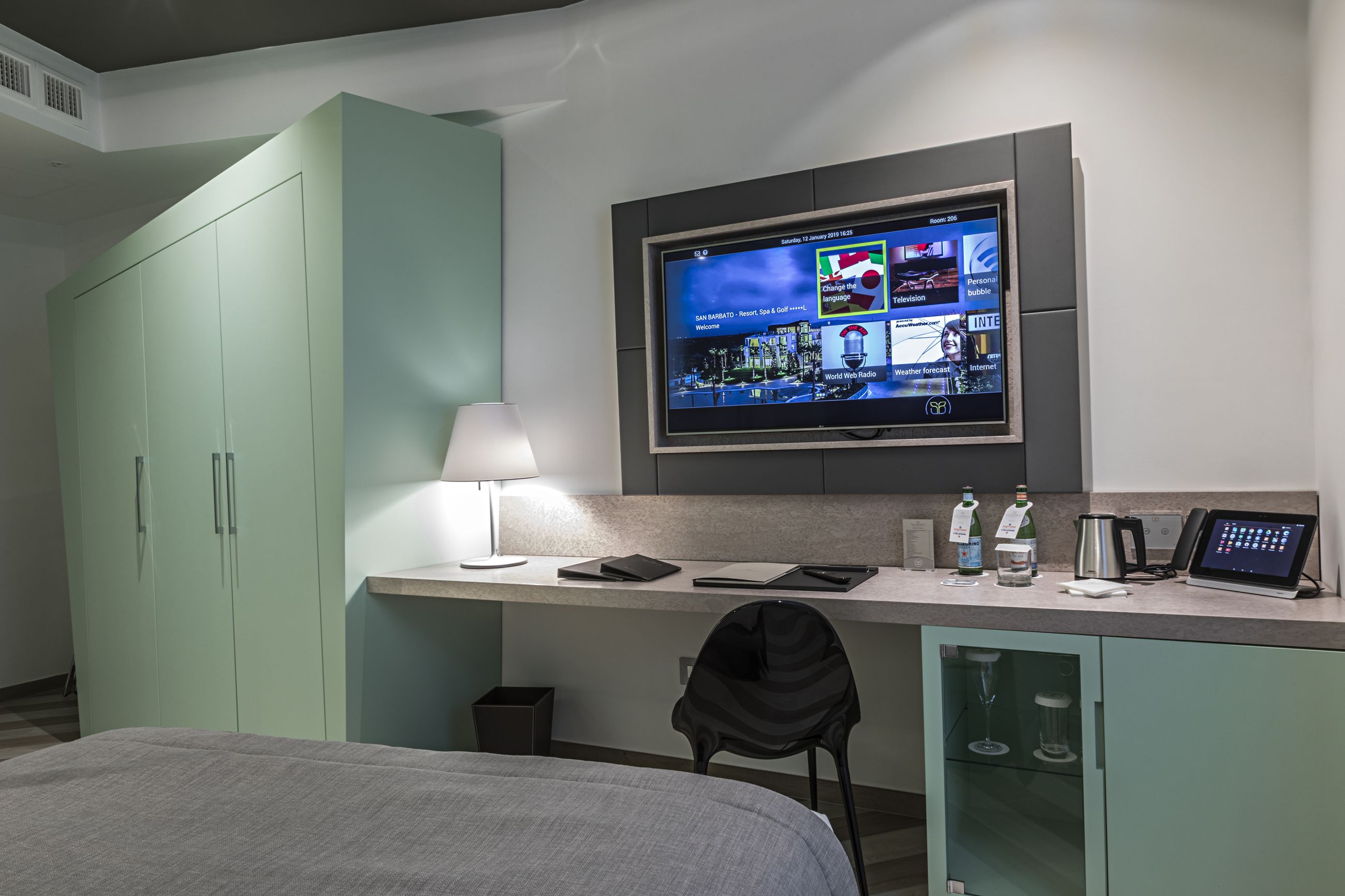 A hotel room with a bed, a desk with a computer, a TV mounted on the wall, and a minibar.