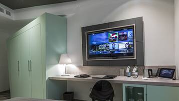 A hotel room with a bed, a desk with a computer, a TV mounted on the wall, and a minibar.