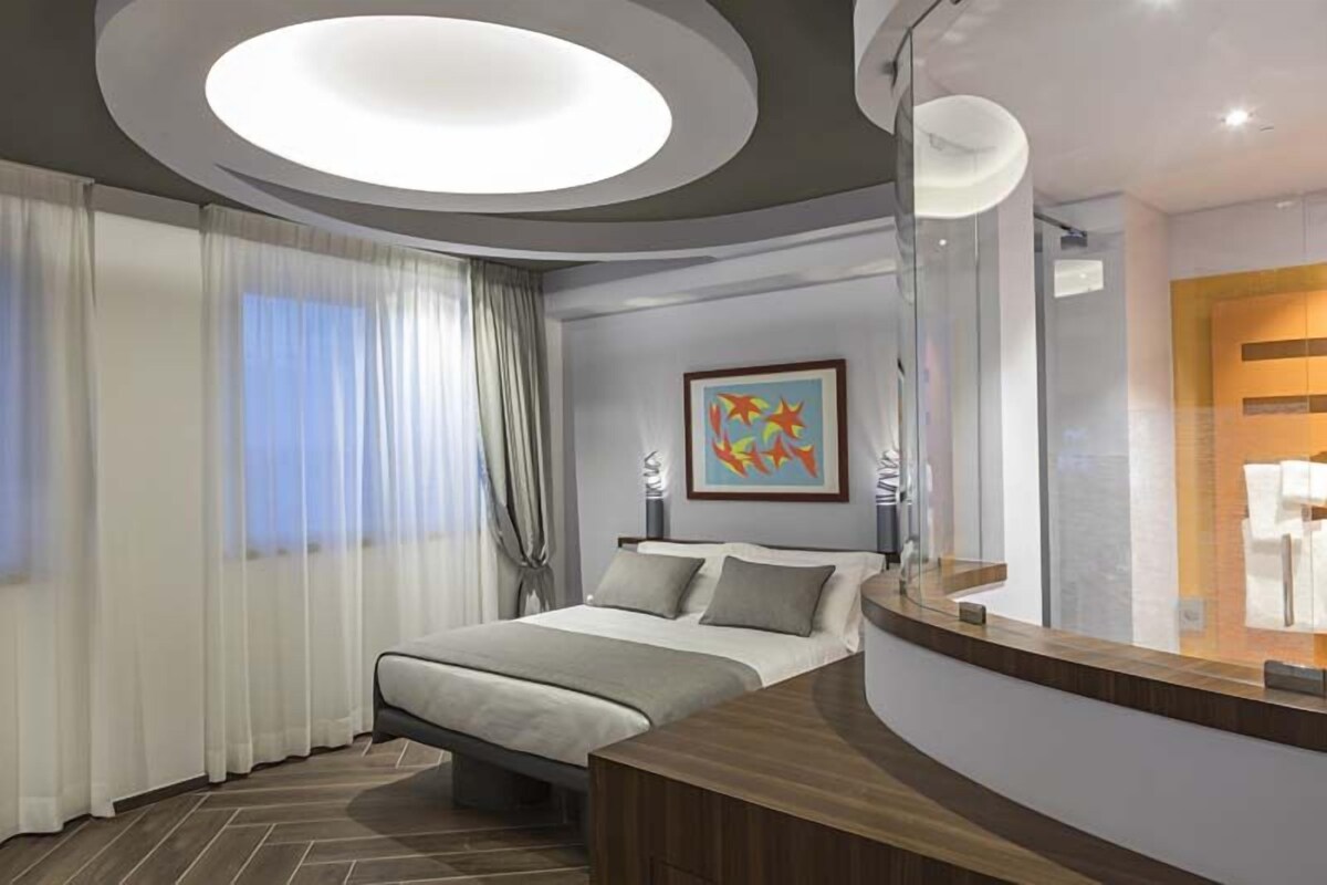 A modern hotel room with a bed, a large mirror, a painting, and a bathroom with a shower and toilet.