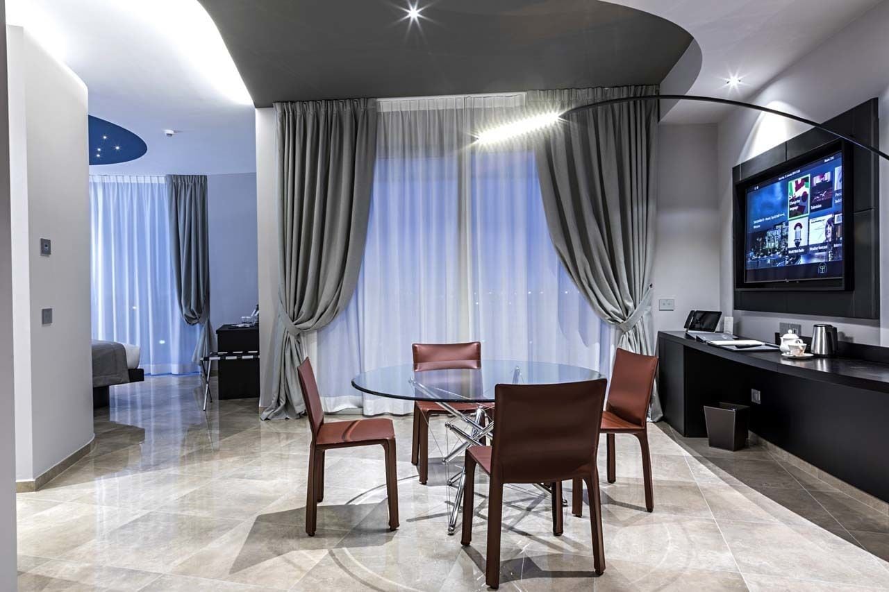 Una habitación de hotel moderna con una mesa redonda de vidrio, sillones de cuero, un televisor de pantalla plana y vistas a la ciudad.