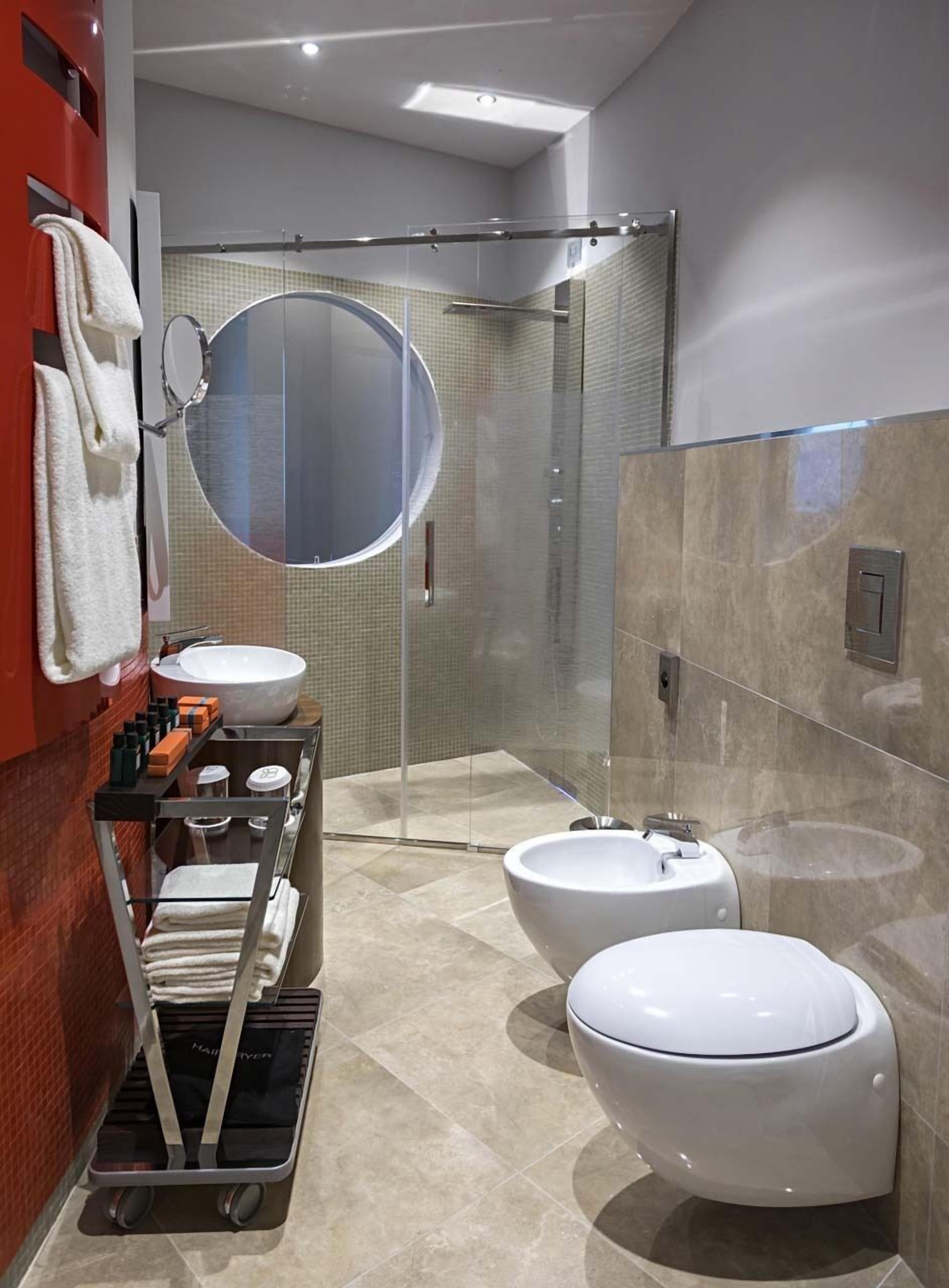 A modern bathroom with a glass-enclosed shower, a round mirror, and two toilets.