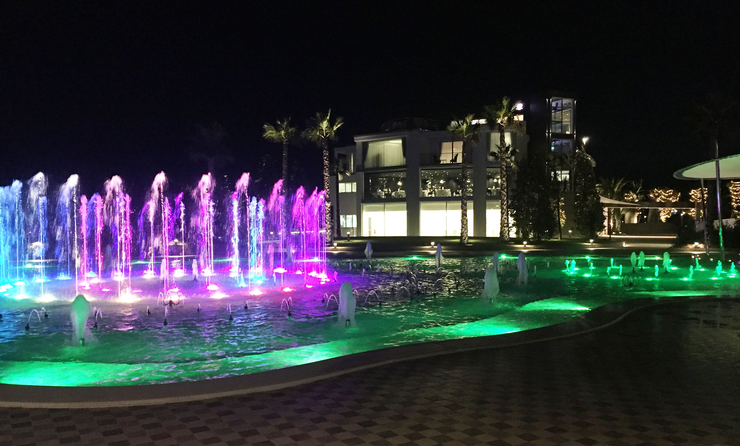 Una fuente iluminada con luces de colores, un edificio moderno con amplios ventanales y palmeras al fondo.
