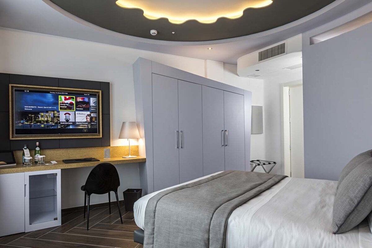 A modern hotel room with a bed, a desk with a computer, a TV mounted on the wall, and grey cabinets.