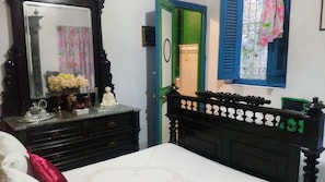 Une chambre à coucher avec un lit, une commode et un miroir. Il y a une porte verte et une fenêtre avec des volets bleus.