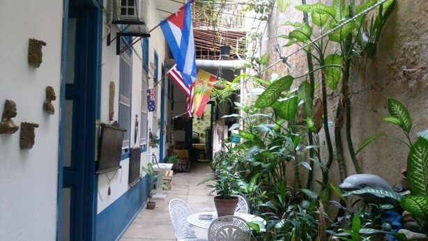 Une ruelle étroite avec un mur blanc, des bordures bleues et une petite table entourée de chaises. Des plantes en pot et un drapeau sont accrochés au mur.