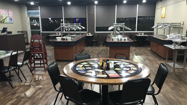 A restaurant with a central round table surrounded by chairs, a bar area with stools, and a view of the city through large windows.