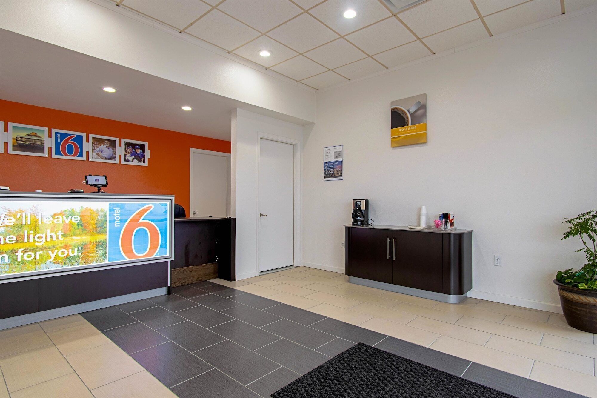 A hotel reception area with a counter, a coffee machine, a potted plant, and a sign advertising the hotel.