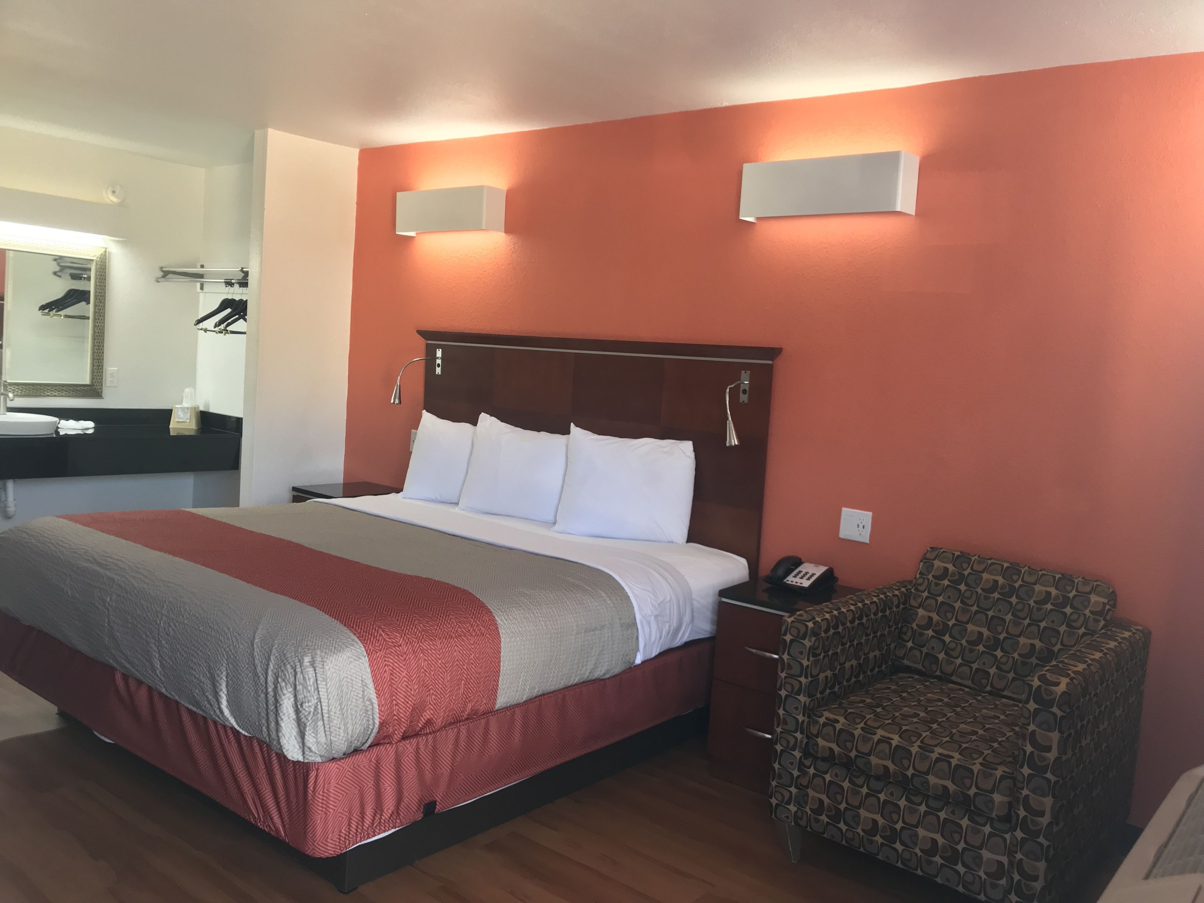 A hotel room with a large bed, a bedside table with a phone, a chair, a mirror, and a bathroom area with a sink and towel rack.