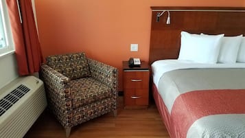 A hotel room with a patterned armchair, a bedside table with a phone, a bed with white linens, and a wall-mounted heater.
