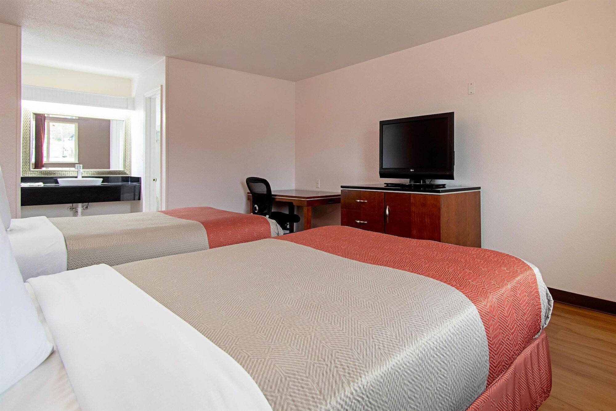 Habitación de hotel con dos camas, un escritorio, una silla, televisión y baño con espejo y lavamanos.