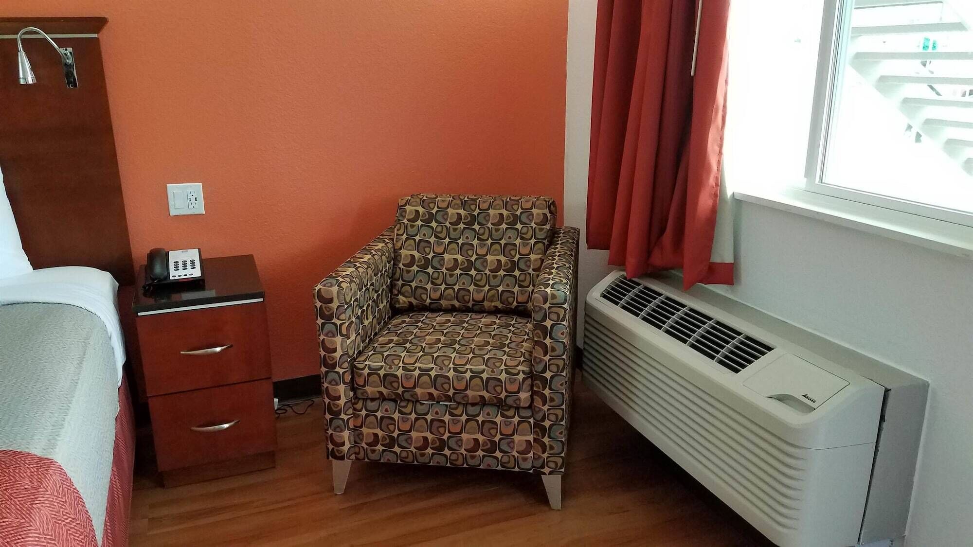 A small, compact room with a patterned armchair, a bedside table with a telephone, a heater, and a window with curtains.