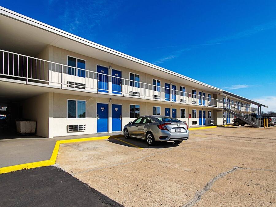 Un motel de dos pisos, con varias habitaciones de puertas azules y un estacionamiento con un coche.
